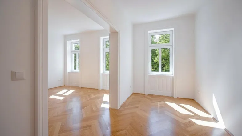 Heller, leerer Raum mit Fischgrätparkettboden und großen Fenstern, die viel Tageslicht hereinlassen.