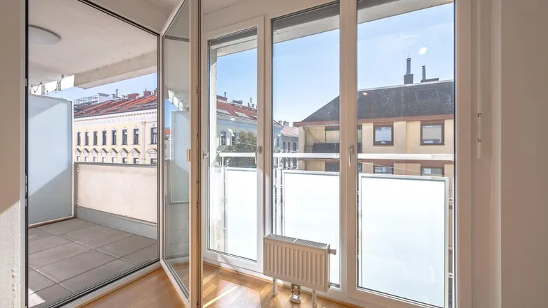 Helles Zimmer mit Parkettboden und Zugang zu einem sonnigen Balkon mit Stadtblick.