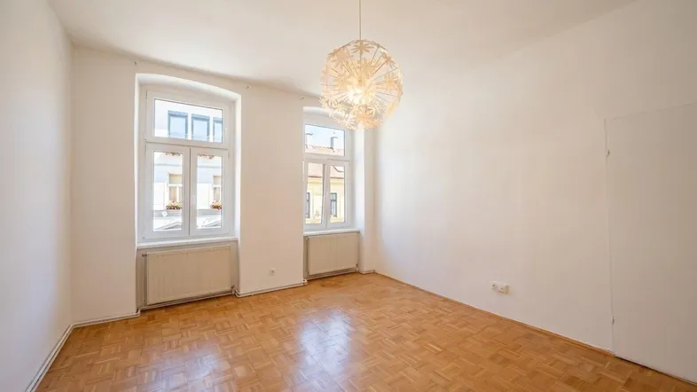 Helles Zimmer mit Fischgrätparkettboden, zwei großen Fenstern und moderner Deckenleuchte.