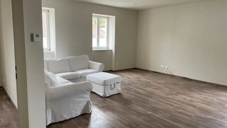 Helles Wohnzimmer mit Holzboden, weißen Wänden und zwei Fenstern, teilweise möbliert.