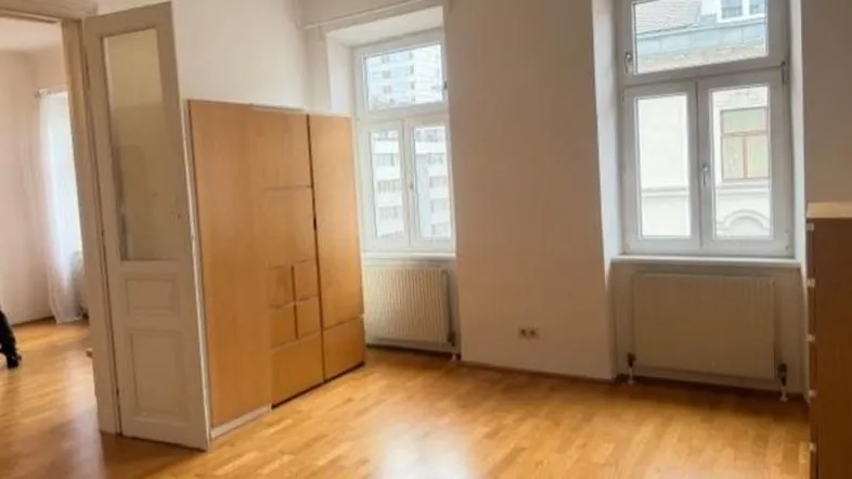 Helles Zimmer mit Parkettboden, hohen Fenstern und einem Kleiderschrank.