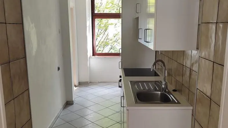 Schmale Küche mit Spüle, Oberschränken und Fenster, ausgestattet mit braunen Fliesen.