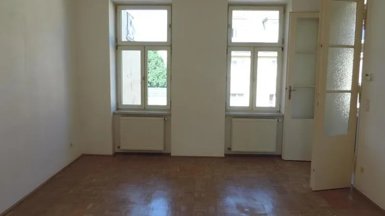Heller Wohnraum mit Parkettboden, zwei großen Fenstern und Heizkörpern.