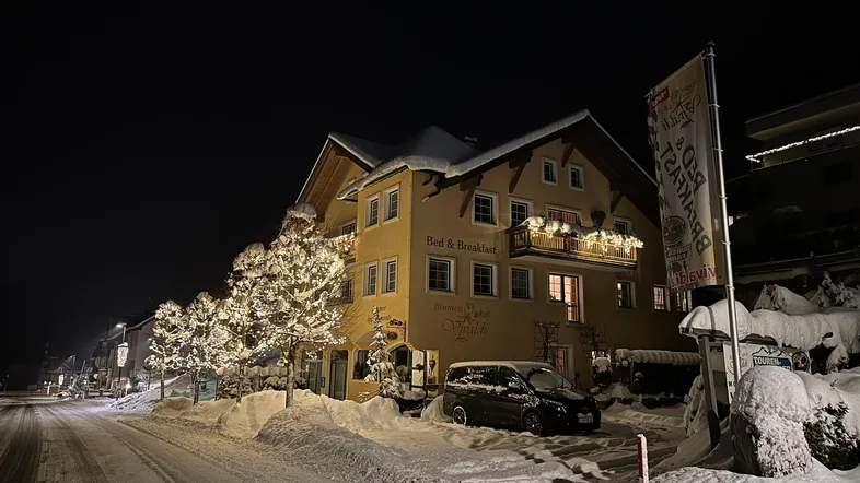 Traditionelles Gebäude bei Nacht im Schnee, festlich beleuchtet mit Lichterketten an Bäumen und einem geparkten Auto.