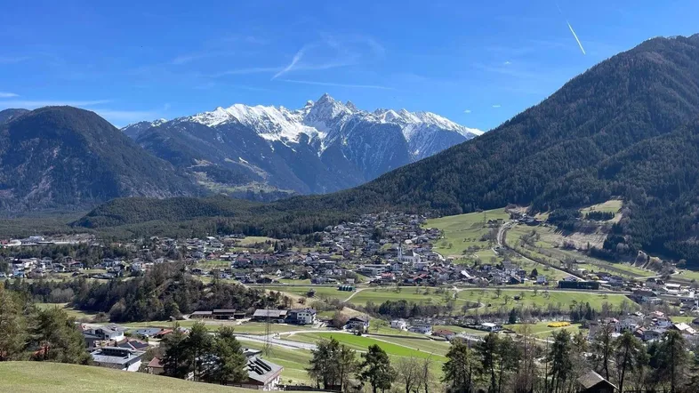 Panoramablick auf ein malerisches Dorf in einem Tal, umgeben von grünen Hügeln und schneebedeckten Bergen.
