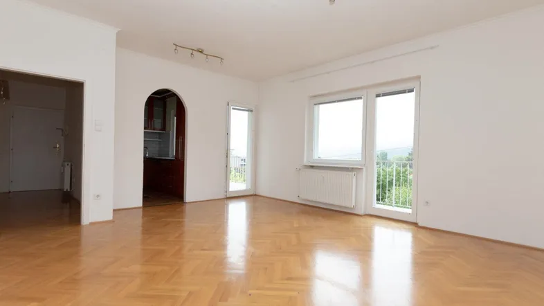 Helles Wohnzimmer mit Fischgrätparkettboden und Zugang zu einem Balkon mit grüner Aussicht.
