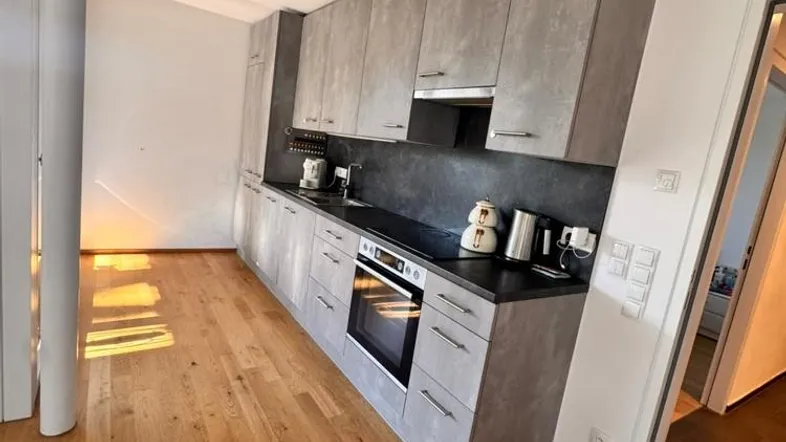 Moderne Küche mit grauen Schränken, dunkler Arbeitsplatte und hellem Holzfußboden, ausgestattet mit Backofen und Kochfeld.