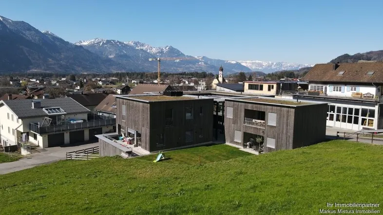 Moderne Gebäude mit Holzfassade und Flachdach, umgeben von grünen Wiesen und Bergpanorama.