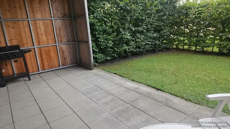 Gepflasterte Terrasse mit Grillbereich und angrenzendem Rasen, umgeben von einer Hecke.