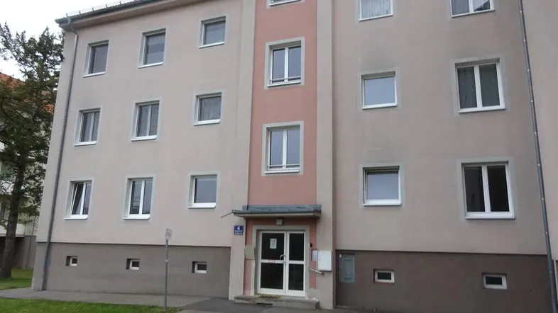 Mehrstöckiges Wohngebäude mit beigefarbener Fassade, rosa Akzenten und gepflegtem Vorgarten.