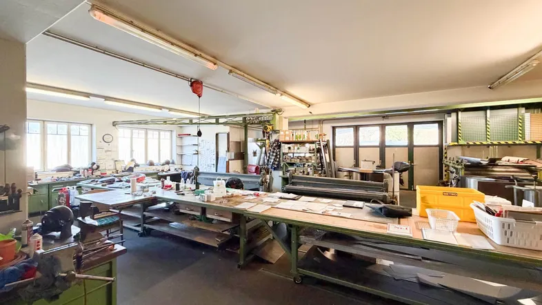 Geräumige Werkstatt mit mehreren Arbeitsbereichen, Maschinen und natürlichem Lichteinfall durch große Fenster.