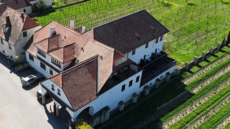 Luftaufnahme eines Gebäudekomplexes mit roten Ziegeldächern, umgeben von grünen Weinbergen unter strahlendem Himmel.