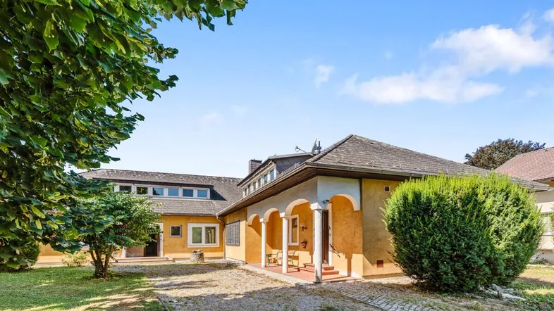 Gelbes Haus mit Satteldach, Kiesauffahrt und gepflegter Gartenanlage unter blauem Himmel.
