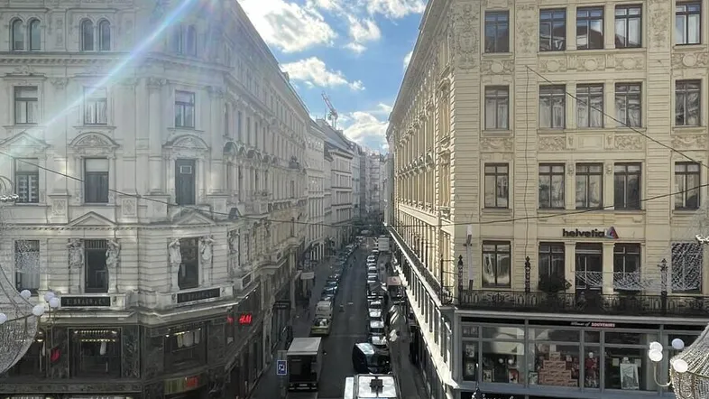 Blick auf eine belebte Stadtstraße mit historischen Gebäuden und Sonnenschein, der die urbane Atmosphäre hervorhebt.