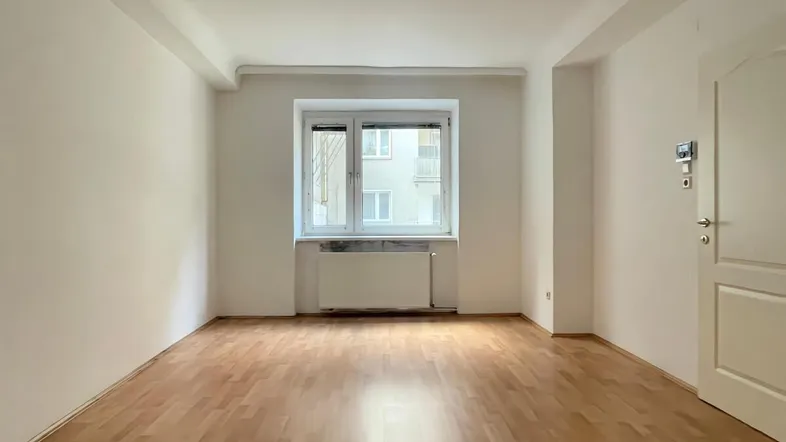 Helles Zimmer mit Parkettboden, weißen Wänden und einem Fenster, das viel Tageslicht hereinlässt.