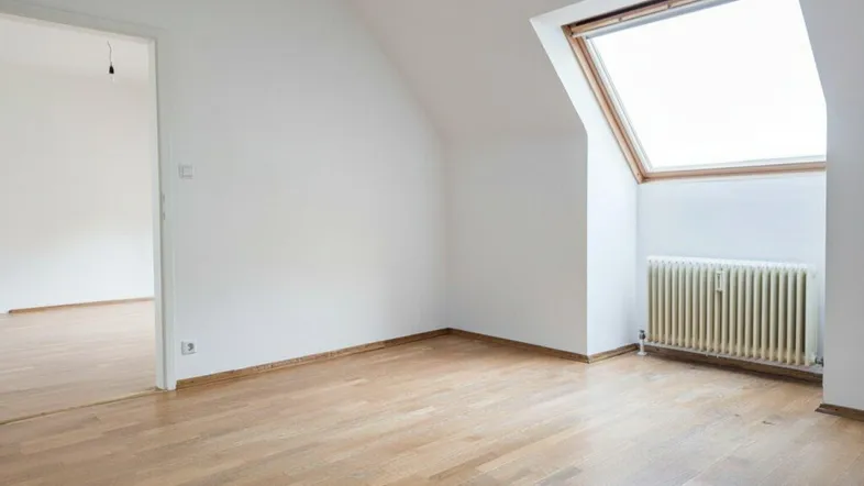 Helles Dachzimmer mit Holzboden, Dachfenster und Heizkörper, bereit zur Einrichtung.