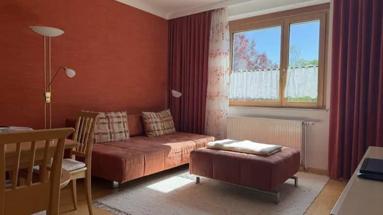 Gemütliches Wohnzimmer mit roter Akzentwand, bequemer Couch und Esstisch am Fenster.