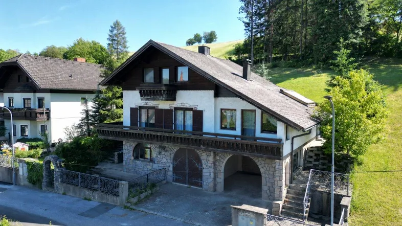 Traditionelles Haus mit Steinfassade und Holzbalkonen, umgeben von grüner Landschaft unter blauem Himmel.