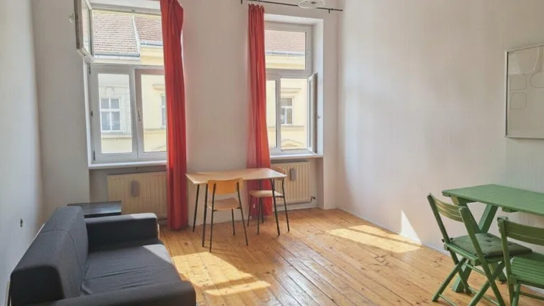 Helles Wohnzimmer mit Holzboden, zwei Fenstern und einfacher Einrichtung.