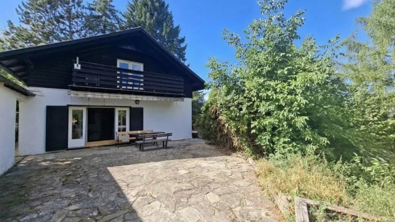 Hausfassade mit gepflasterter Terrasse und Sitzgelegenheiten im Freien unter einem Balkon.