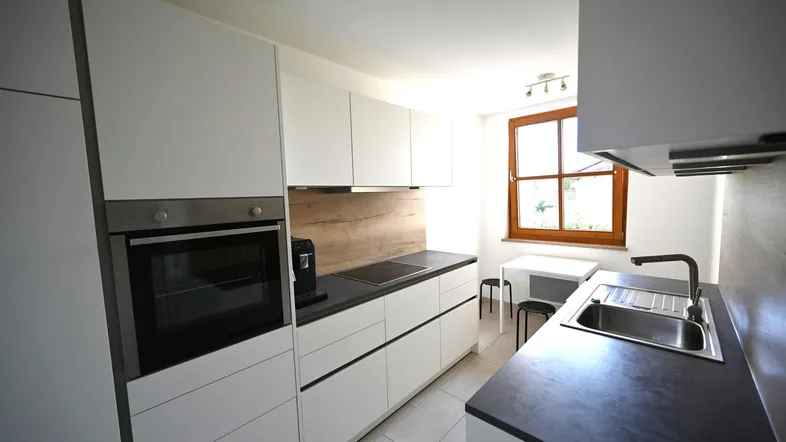Moderne Küche mit weißen Schränken, dunkler Arbeitsplatte, Einbauofen und Fenster für Tageslicht.