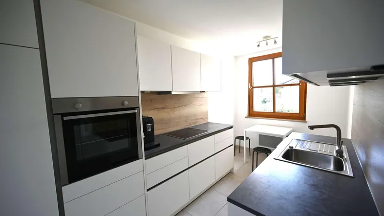 Moderne Küche mit weißen Schränken, dunkler Arbeitsplatte, integriertem Backofen und Fenster.