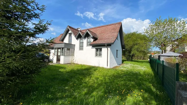 Weißes Haus mit rotem Ziegeldach und Gauben, umgeben von einer grünen Wiese unter blauem Himmel.