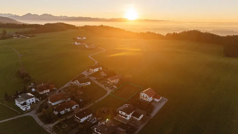Luftaufnahme eines ländlichen Wohngebiets mit grünen Feldern und Häusern bei Sonnenuntergang, umgeben von Bergen.