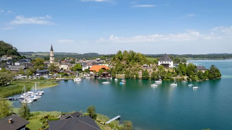 Panoramablick auf einen malerischen See mit Segelbooten, einer charmanten Stadt und umliegenden Häusern unter blauem Himmel.