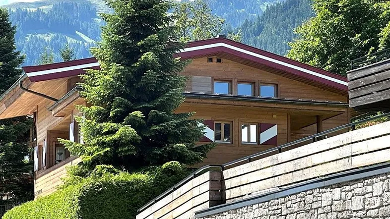 Chalet-Stil Haus mit Holzfassade und Satteldach, umgeben von grünen Bäumen und Bergen.