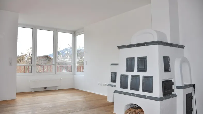 Helles Wohnzimmer mit traditionellem Kachelofen, Holzboden und großen Fenstern mit Blick ins Grüne.