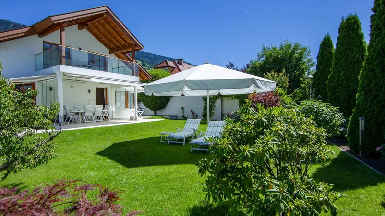 Großzügiger Gartenbereich mit Rasenfläche, Sonnenliegen und einer überdachten Terrasse am Haus.