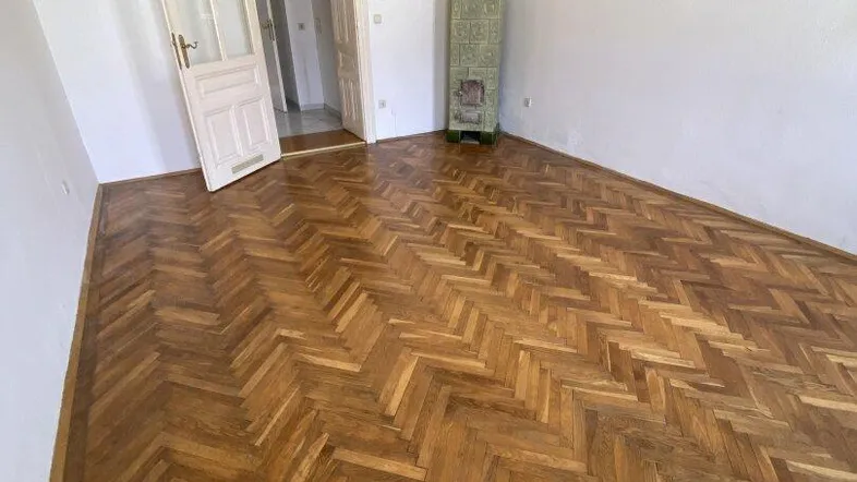 Geräumiges Zimmer mit Fischgrätparkett und traditionellem Kachelofen, ideal als Wohn- oder Schlafzimmer.