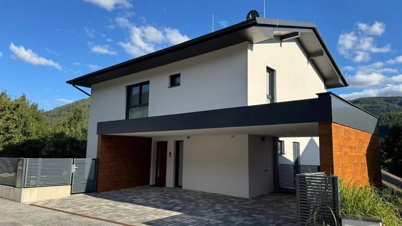 Modernes Einfamilienhaus mit Carport und gepflasterter Auffahrt vor bergiger Kulisse.