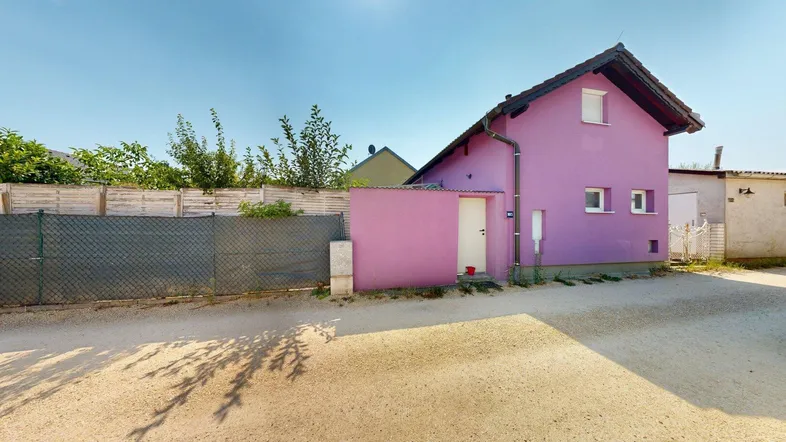 Hausfassade in Lila mit angrenzendem Schuppen und Kiesfläche unter blauem Himmel.