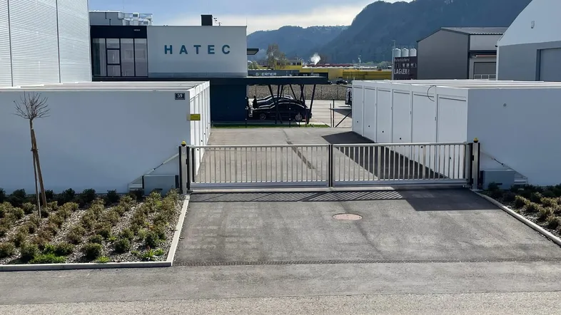 Einfahrt zu einem Gewerbeobjekt mit Schiebetor, umgeben von modernen Gebäuden und Bergen im Hintergrund.