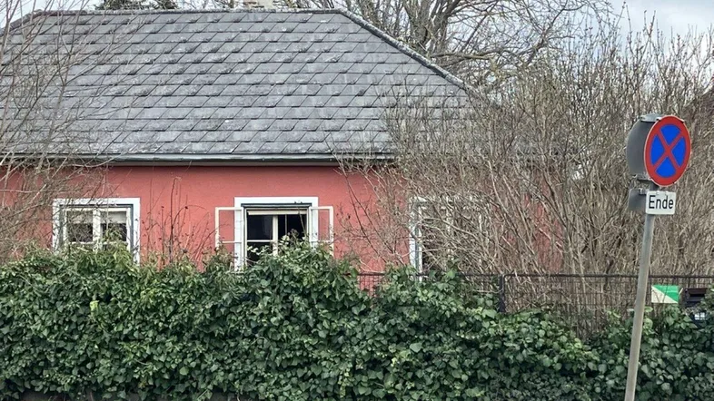 Rotes Haus mit grauer Dachpfanne und grüner Hecke im Vorgarten an einer Straße.