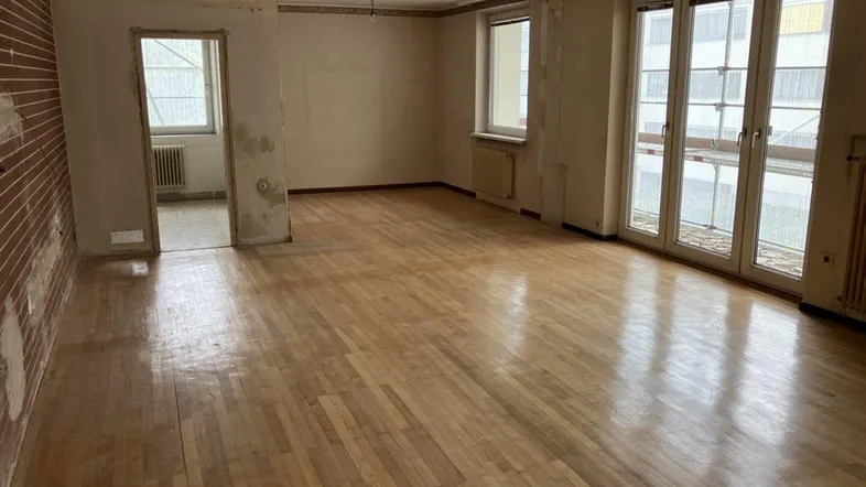 Geräumiges Wohnzimmer mit Holzboden und großen Fenstern, bereit für eine Modernisierung.