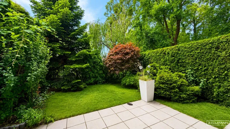 Gepflegter Garten mit Rasenfläche, hohen Hecken und einer kleinen gepflasterten Terrasse.