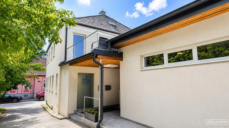 Modernes Gebäude mit hellem Putz und einem überdachten Eingangsbereich unter blauem Himmel.