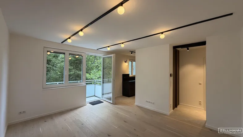 Helles Wohnzimmer mit modernem Schienensystem an der Decke und Zugang zum Balkon mit Grünblick.