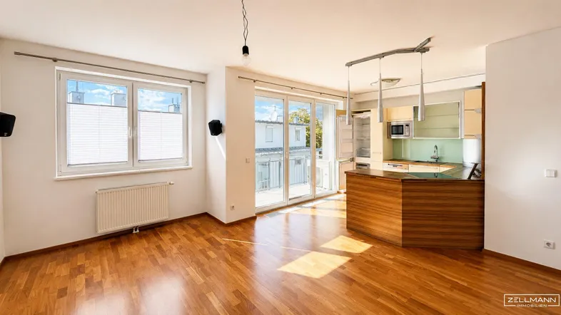 Heller offener Wohnbereich mit Holzboden, großen Fenstern und integrierter Küche mit Balkonzugang.