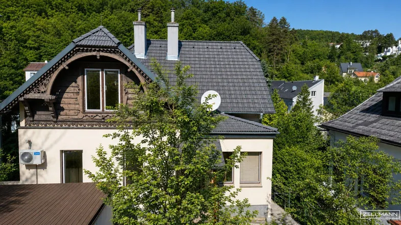 Hausfassade mit traditionellen Holzelementen, dunklem Ziegeldach und umgeben von üppigem Grün.