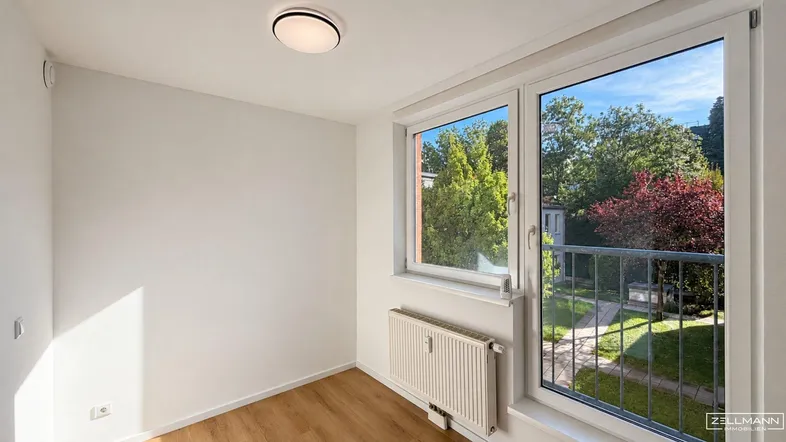 Helles Zimmer mit Holzboden, weißen Wänden und Zugang zu einem kleinen Balkon mit Grünblick.