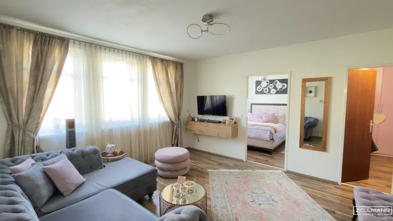 Helles Wohnzimmer mit gemütlichem Sofa, Holzboden und Blick in ein angrenzendes Schlafzimmer.
