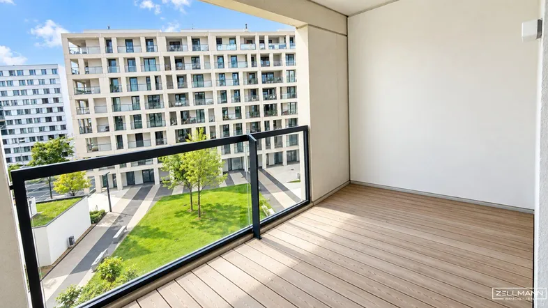 Geräumiger Balkon mit Holzbelag und Glasgeländer, bietet Blick auf den begrünten Innenhof und moderne Gebäude.