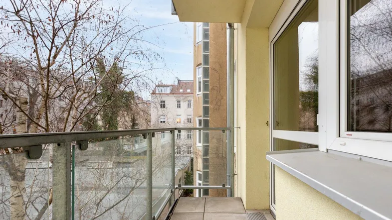 Balkon mit Glasgeländer und Blick auf die umliegenden Bäume und Gebäude der Stadt.