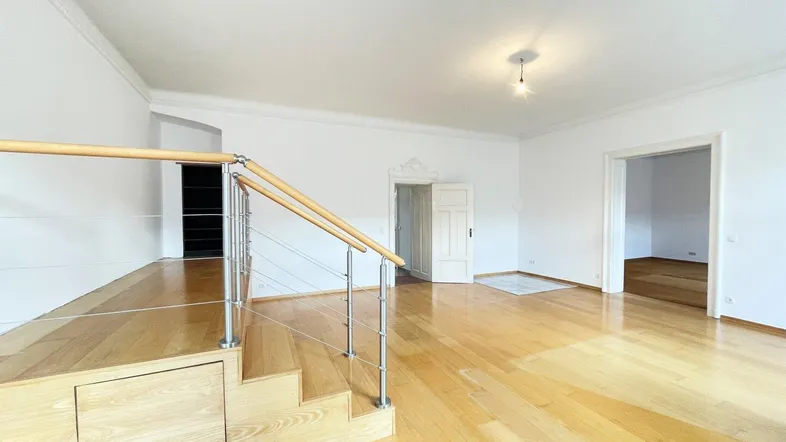 Geräumiges Wohnzimmer mit hellem Parkettboden, hohen Decken und einer kleinen Treppe zu einer erhöhten Ebene.
