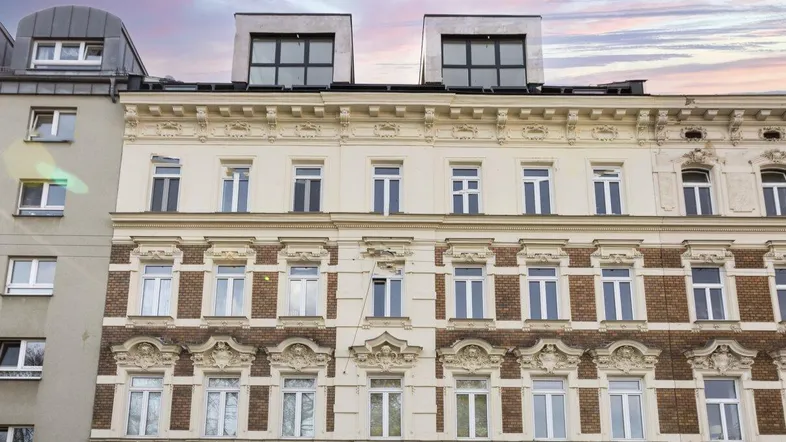 Historische Altbaufassade mit modernem Dachausbau und großen Fenstern unter einem dramatischen Himmel.