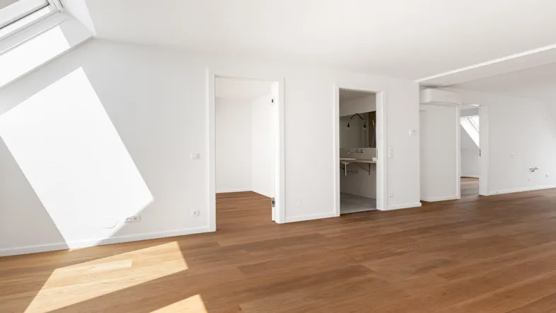 Heller, leerer Raum mit Holzboden und Dachfenstern, der zu einem modernen Badezimmer führt.
